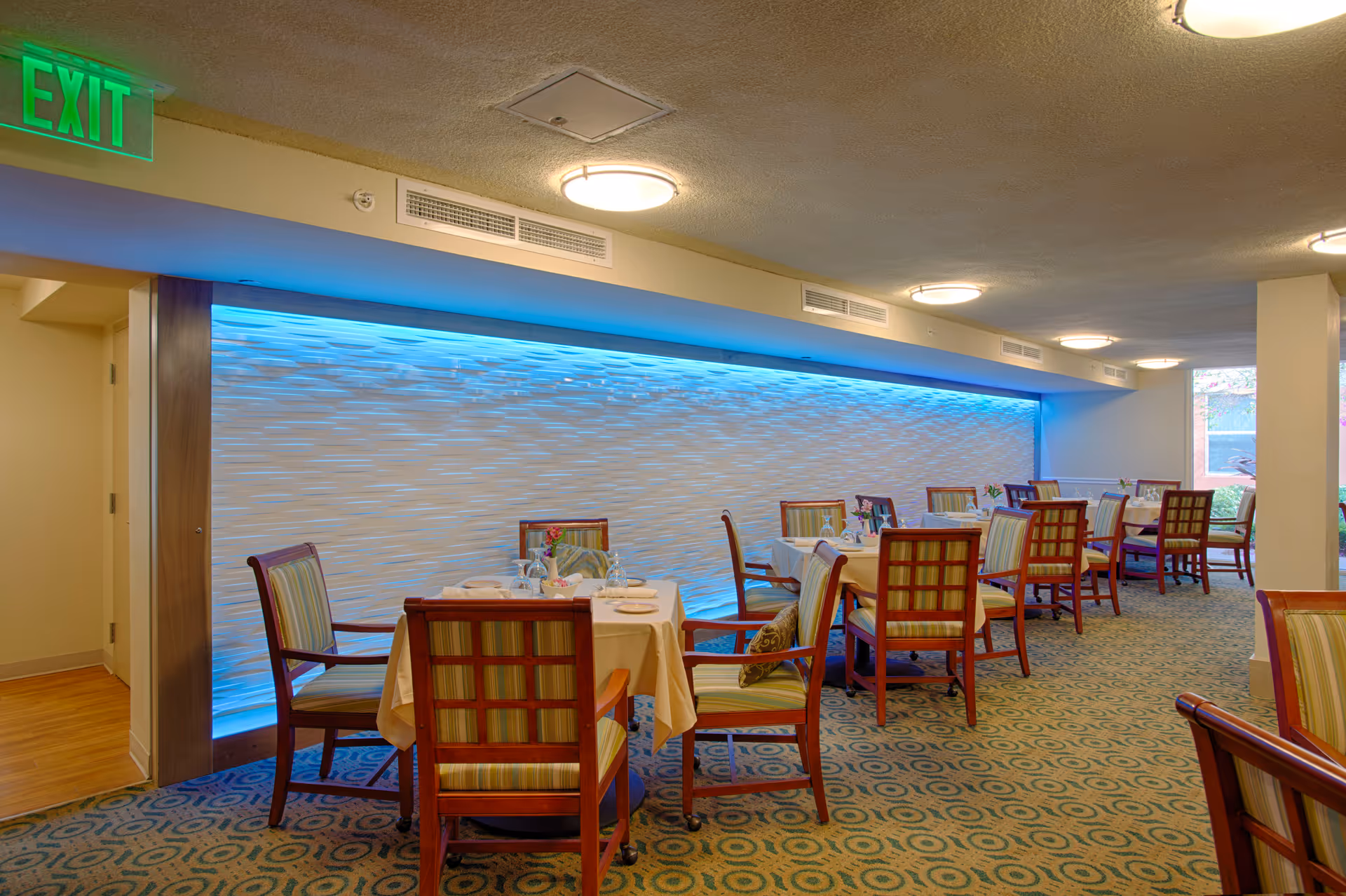 Dining room with multiple set tables and chairs facing a blue-lit textured wall under ceiling lights.