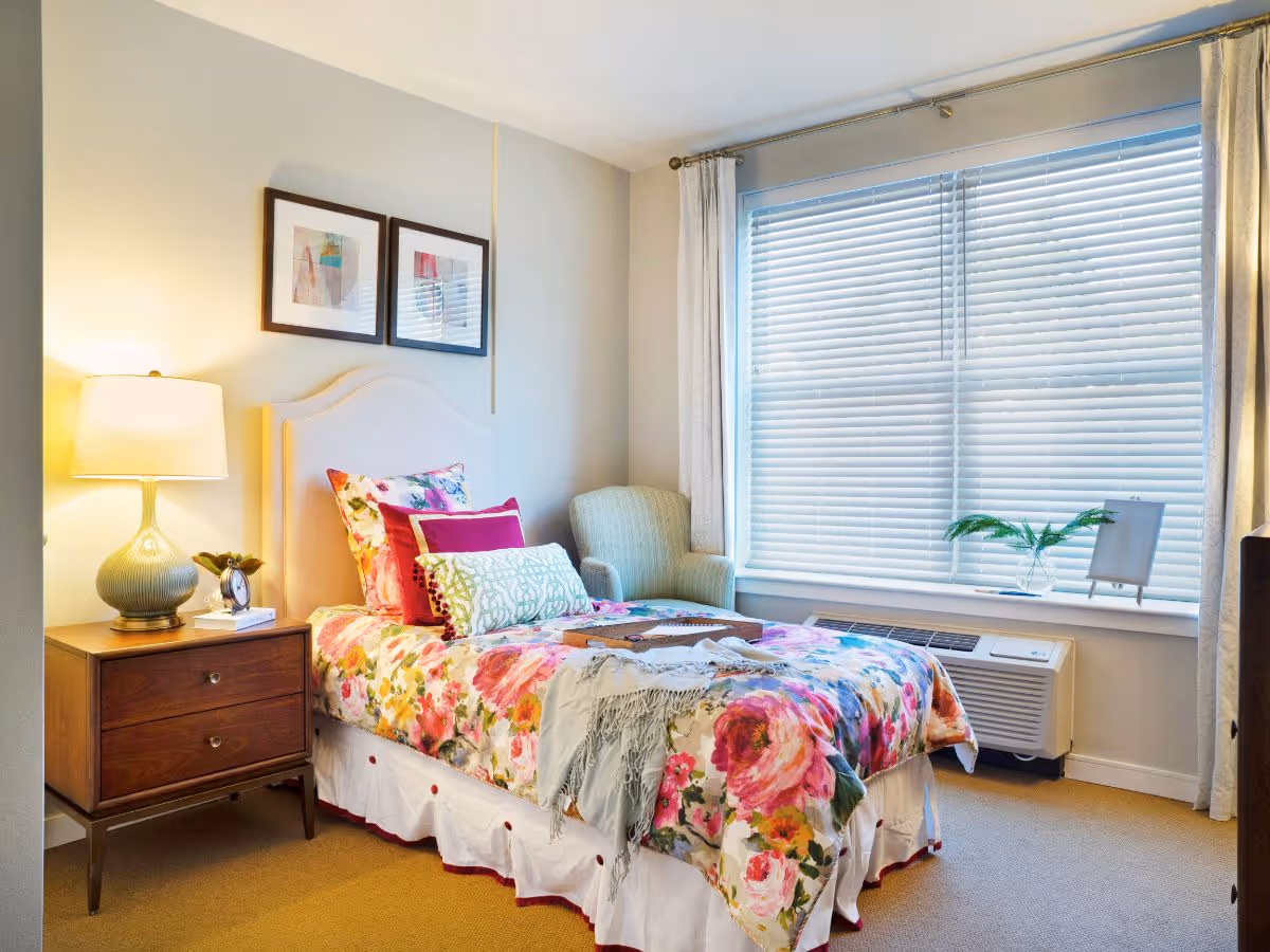 Sunny bedroom with a floral-covered single bed, wooden nightstand and lamp, upholstered chair, and a large window with blinds.