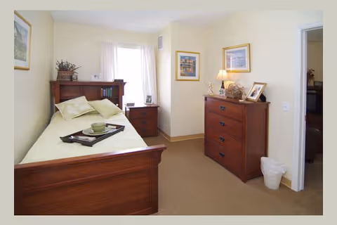 A cozy bedroom with a wooden bed featuring light-colored bedding and pillows. A tray with a cup and plate is placed on the bed. There is a wooden nightstand with a lamp and picture frame next to the bed, and a wooden dresser with a lamp, framed pictures, and decorative items against the wall. The room has light-colored walls, a window with sheer curtains, and framed artwork hanging on the walls.