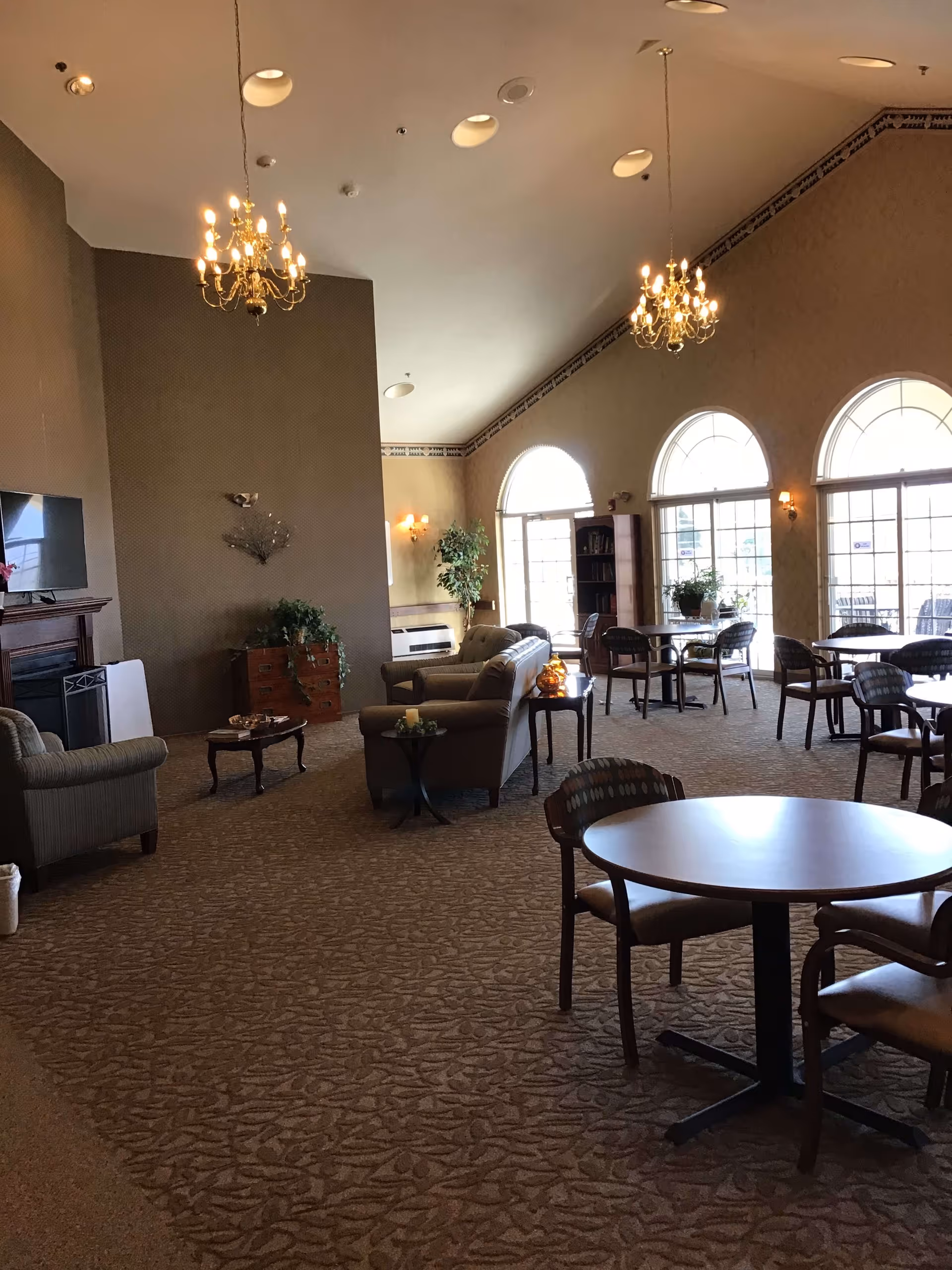 A spacious senior living common area with carpeted floor, multiple round tables with chairs, sofas, and armchairs. The room features high ceilings with two chandeliers, large arched windows letting in natural light, and decorative plants. There is a fireplace with a TV mounted above it on one wall.
