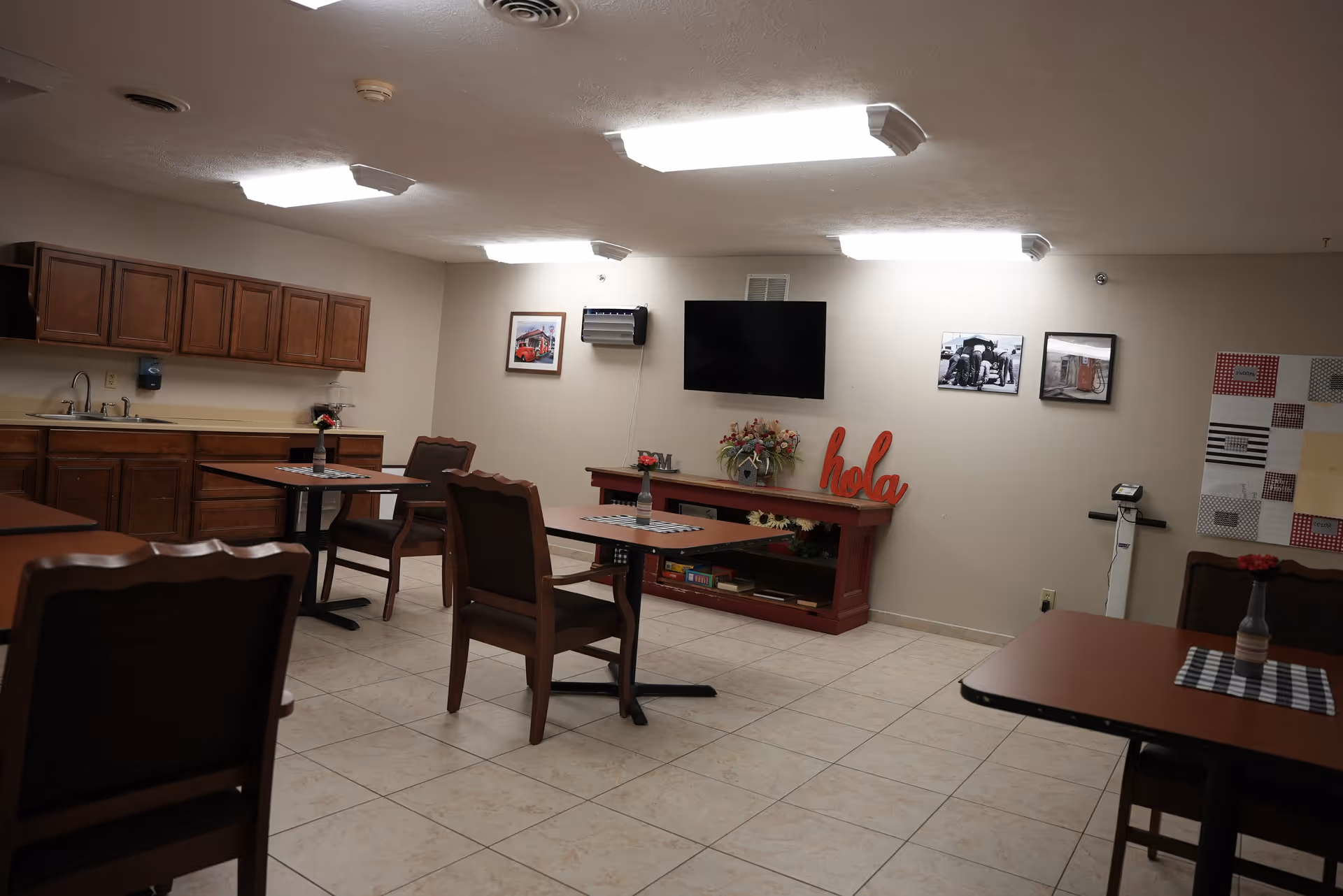 Community dining/activity room with tables and chairs, a kitchenette along one wall, a wall-mounted TV, and a decorative shelf with a 'hola' sign.