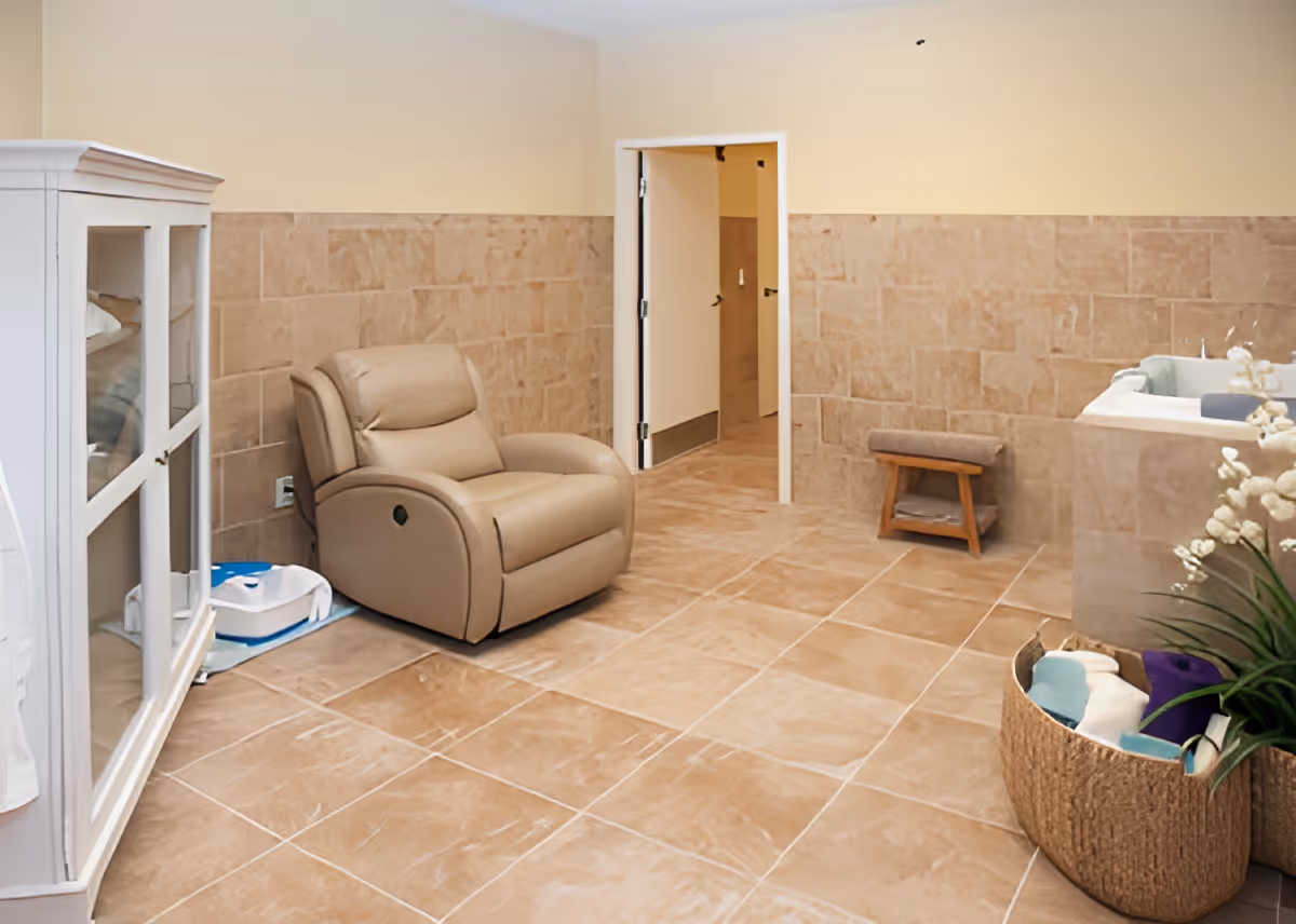 A spacious room with beige tiled floors and walls halfway up, featuring a comfortable beige recliner chair, a white cabinet with glass doors, a small wooden stool, and a large built-in bathtub. There is a basket with rolled towels and a plant near the bathtub. An open door leads to another room with similar tiling.