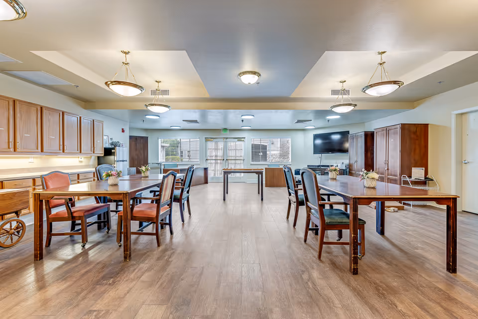 Large communal dining/activity room with wooden tables and chairs, overhead lights, cabinets along the left wall, and a wall-mounted TV at the far end.