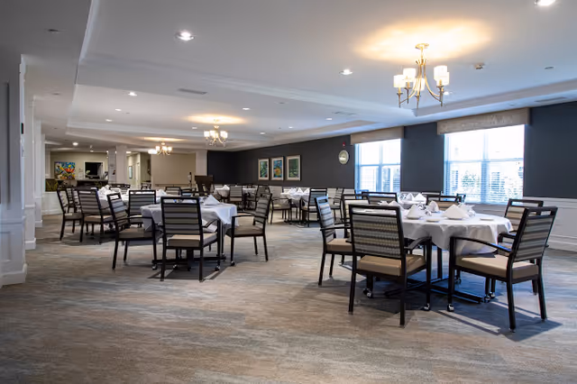 A spacious dining room with multiple round tables covered with white tablecloths and neatly arranged chairs. The room has large windows allowing natural light, chandeliers on the ceiling, and framed artwork on the walls.