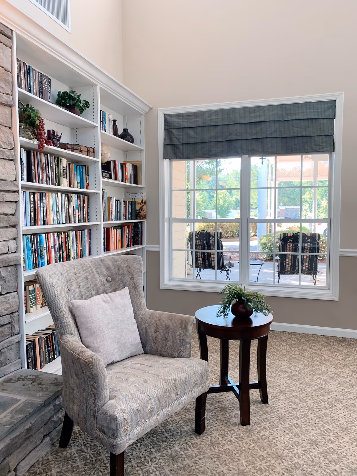 A cozy reading nook featuring a cushioned armchair with a pillow, a small round wooden side table with a decorative plant, a built-in white bookshelf filled with books and decorative items, and a large window with a gray Roman shade overlooking an outdoor patio with chairs and greenery.
