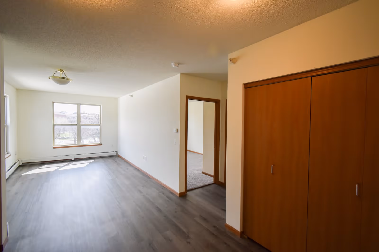 Empty room in a senior apartment with light-colored walls, large window letting in natural light, wood laminate flooring, and a wooden closet with sliding doors. A doorway leads to another room with carpeted flooring.