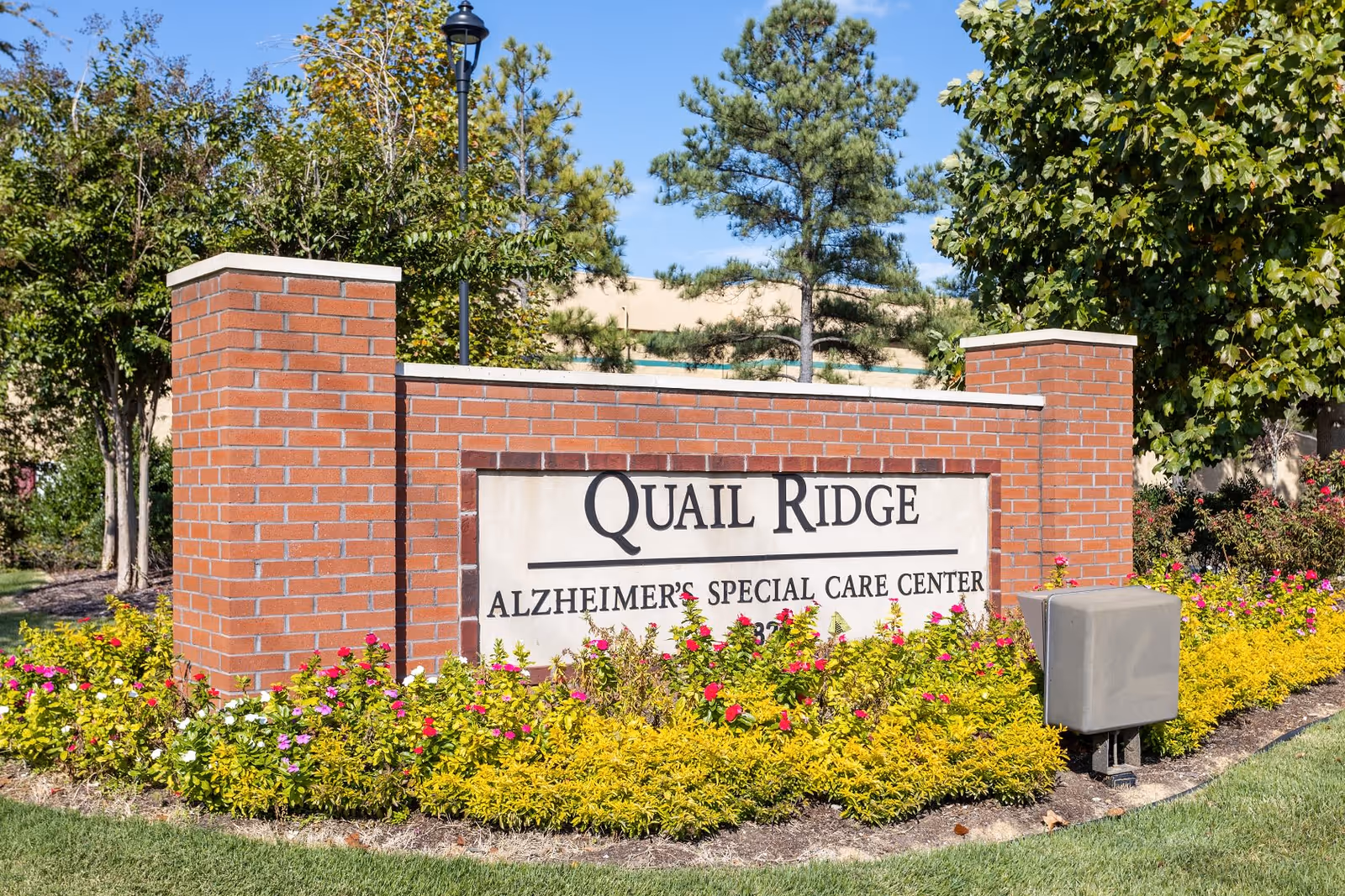 A brick sign for Quail Ridge Alzheimer's Special Care Center surrounded by colorful flowers and greenery with trees and a clear blue sky in the background.