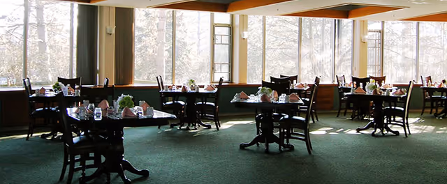 A dining room with multiple dark wooden tables and chairs arranged neatly. Each table is set with folded napkins, glasses, and small floral centerpieces. Large windows allow natural light to fill the room, showing a view of trees outside.