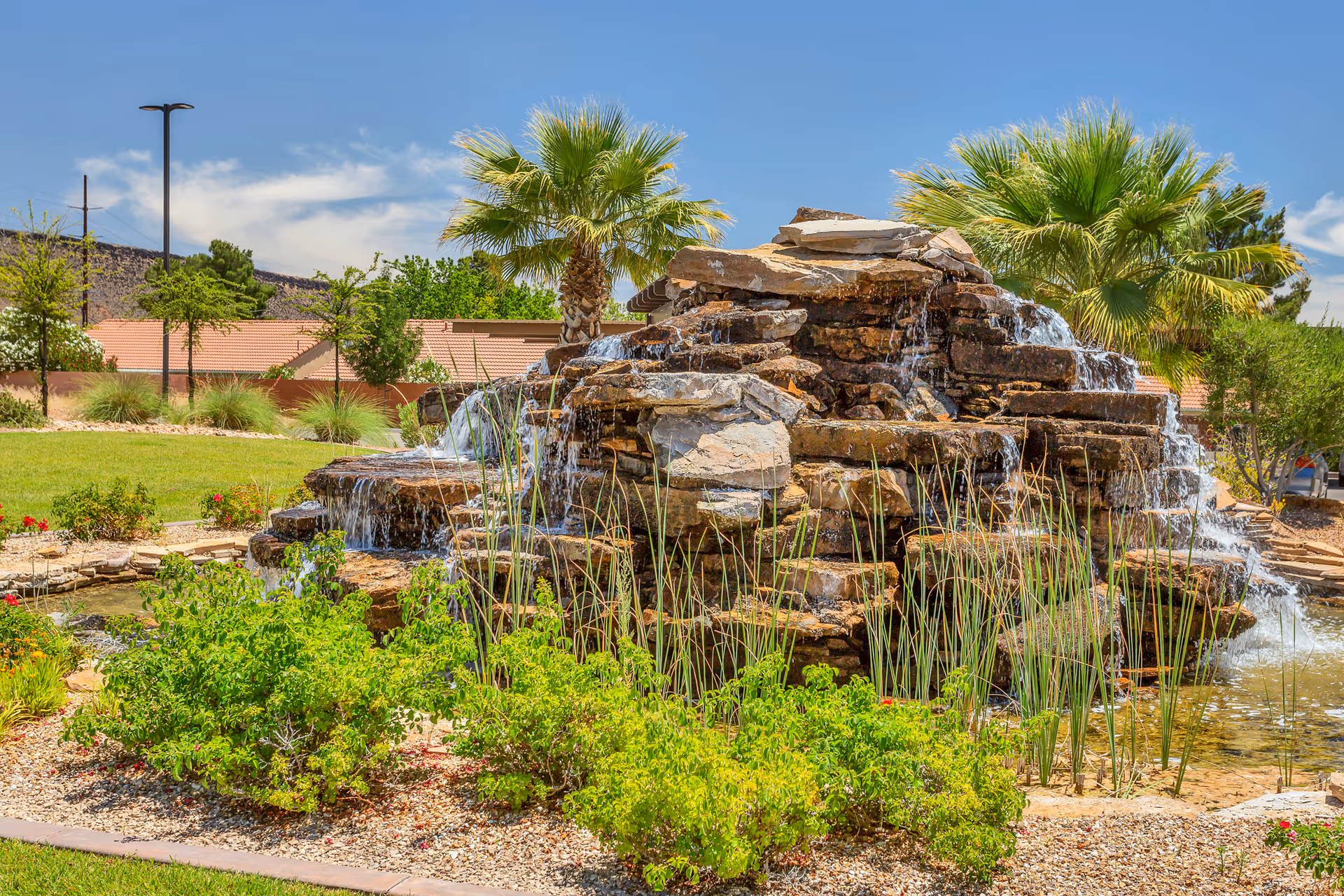 Rock waterfall and pond surrounded by landscaping and palm trees under a sunny blue sky.