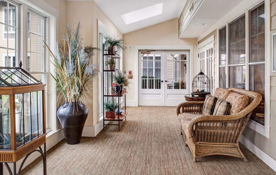 A bright and cozy indoor sitting area with large windows on the left letting in natural light. The space features a wicker loveseat with patterned cushions and pillows, a small round wooden side table with a decorative birdcage, a tall black vase with ornamental grasses, and a metal shelving unit with potted plants. The floor is carpeted, and there are double glass doors at the far end of the room.