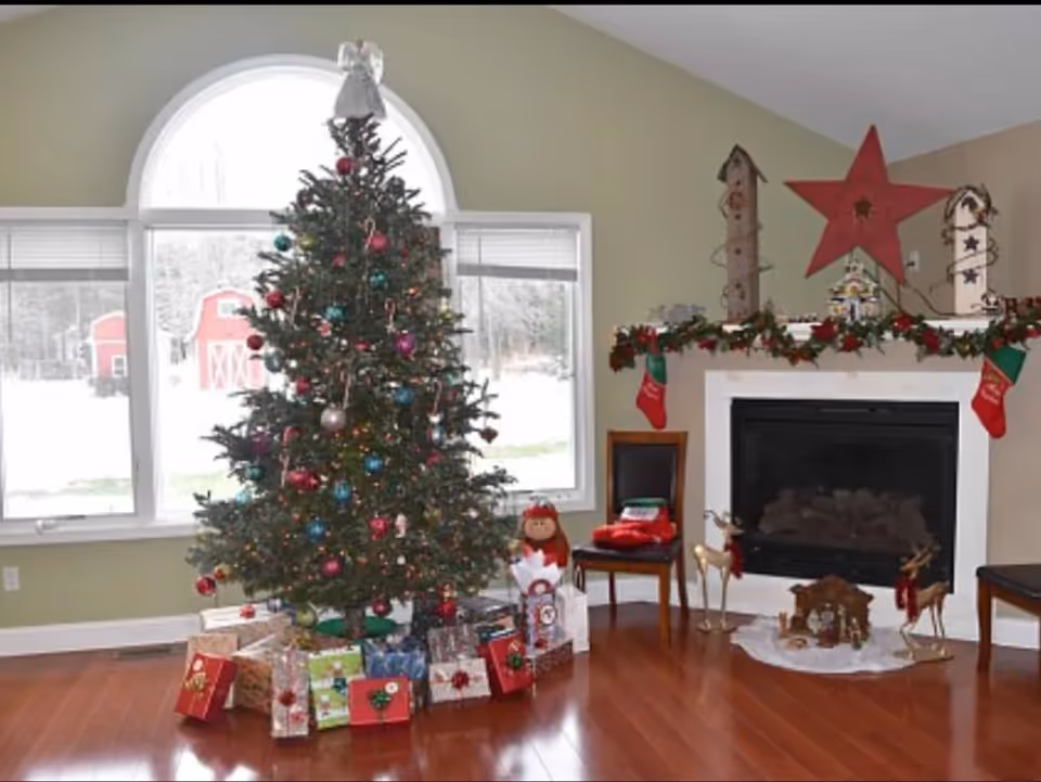 A decorated Christmas tree with colorful ornaments and an angel on top stands in front of a large arched window. Wrapped presents are placed under the tree. To the right, there is a fireplace mantle adorned with garland, stockings, a large red star, and decorative birdhouses. Two chairs and reindeer figurines are also visible near the fireplace.