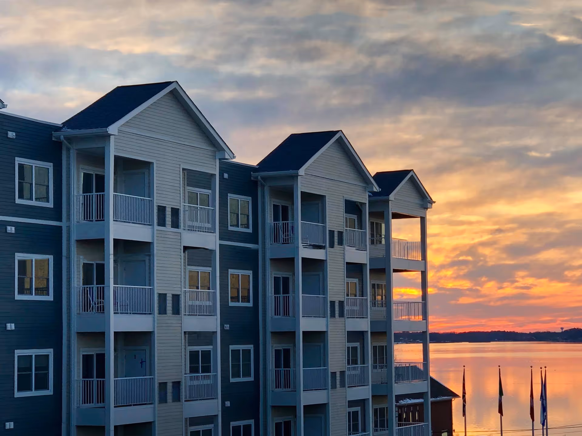 Exterior of a multi-story lakeside residential building with stacked balconies under a colorful sunset sky.