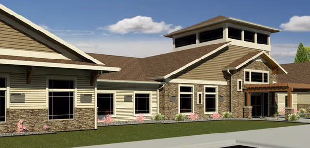 Exterior view of a senior living facility building with beige siding, stone accents, multiple windows, and a brown roof under a partly cloudy sky.