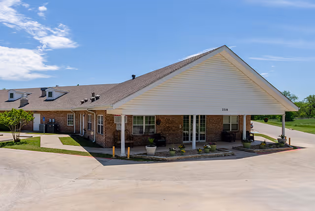 Exterior view of Wheeler Terrace facility showing a single-story brick building with a large covered entrance, potted plants, and a paved driveway under a blue sky with some clouds.