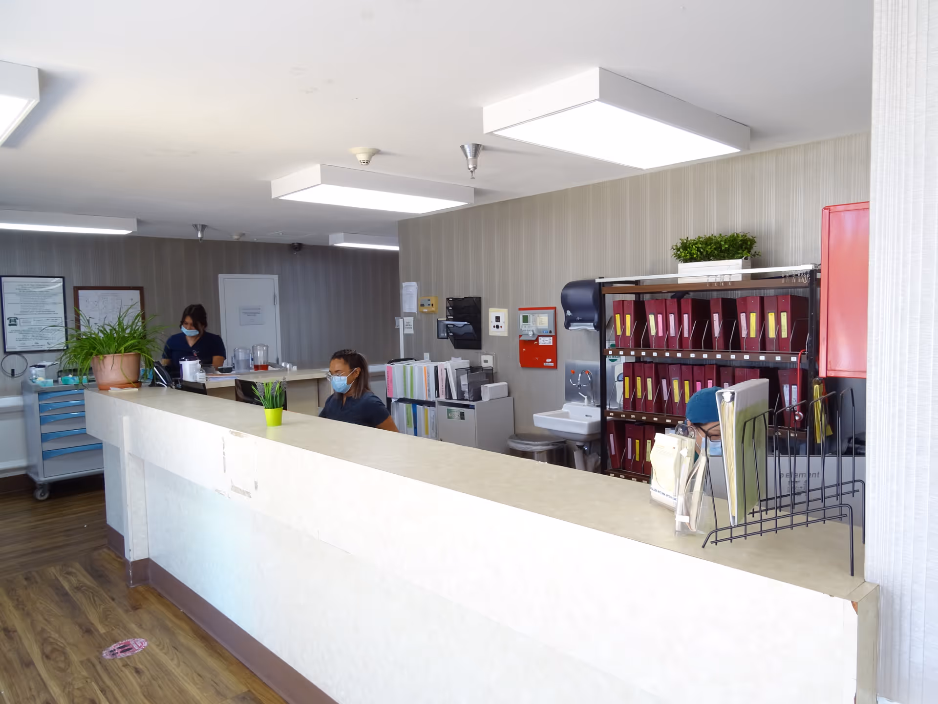 Reception area inside Rosecrans Care Center with two staff members wearing masks seated behind a long counter. The room has wood flooring, overhead fluorescent lighting, office supplies, filing shelves with red binders, a small sink, and potted plants on the counter.