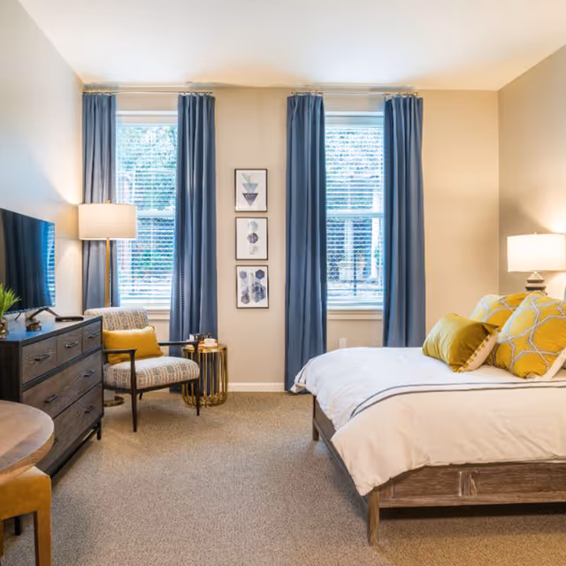 A bright and cozy bedroom featuring a bed with white bedding and yellow accent pillows, two windows with blue curtains, a wooden dresser with a TV on top, a patterned armchair with a yellow pillow, a small round side table, and two lamps providing warm lighting.
