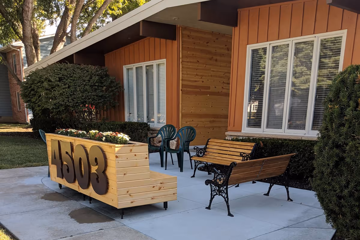 Outdoor patio area with a wooden planter box displaying the number 4503, surrounded by green plastic chairs and wooden benches with decorative black metal armrests. The patio is adjacent to a building with orange and natural wood siding and white-framed windows. There are bushes and trees around the patio.