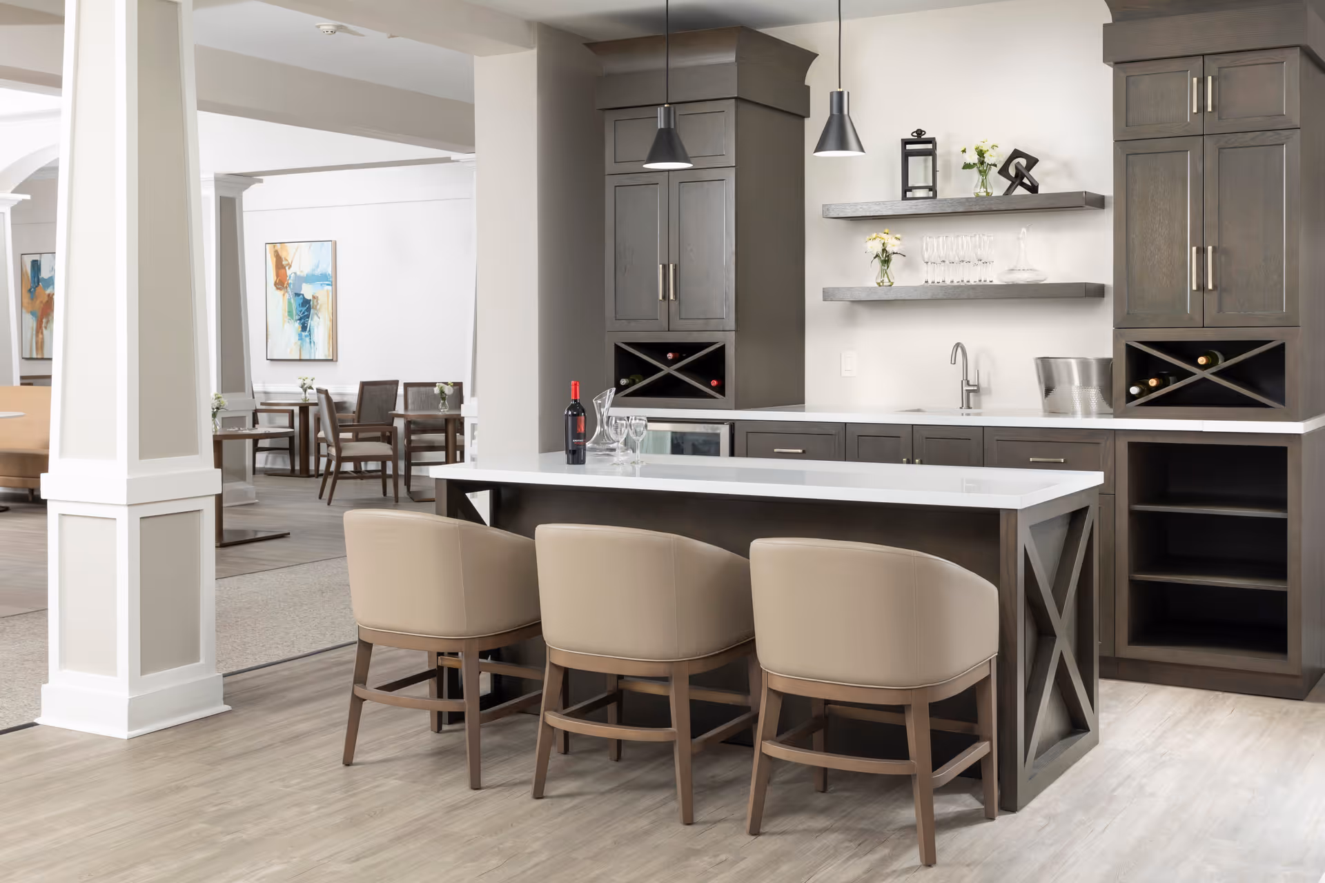 Modern kitchen area with a large island featuring a white countertop and three beige cushioned chairs. The back wall has dark wood cabinets, open shelves with glassware and decorative items, a small sink, and wine storage. In the background, there is a dining area with tables and chairs, and abstract artwork on the walls.