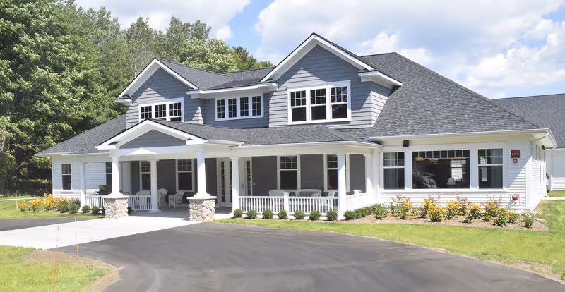 Front exterior view of a large, two-story residential-style building with gray siding and white trim, featuring multiple windows, a covered porch with white railings and stone pillars, surrounded by green lawn and trees under a partly cloudy sky.