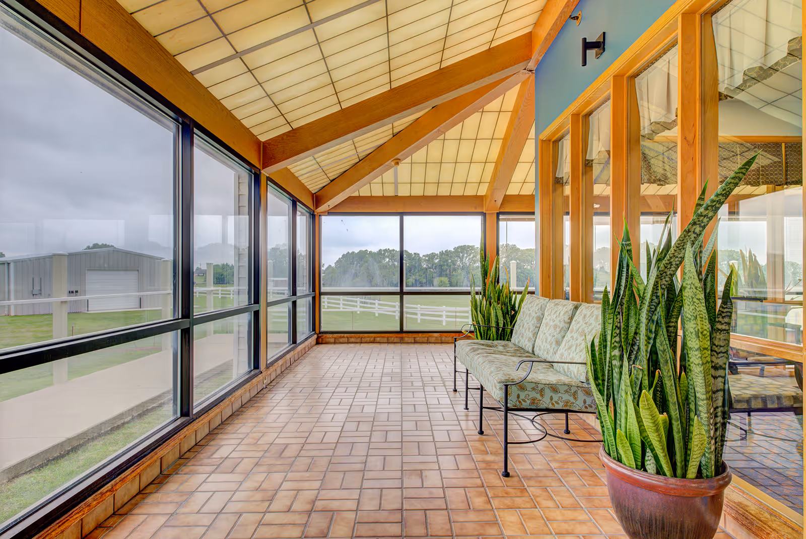 Bright enclosed sunroom with large windows, tiled floor, bench seating and potted plants overlooking a grassy yard.