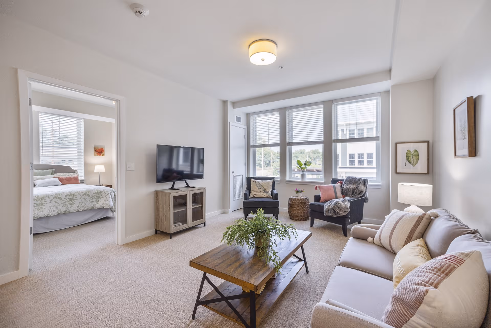 A bright and cozy living room with a beige sofa adorned with multiple pillows, a wooden coffee table with a green plant centerpiece, two dark armchairs with cushions and a throw blanket, a small round side table, and a flat-screen TV on a wooden stand. Large windows let in natural light, and a bedroom with a bed and bedside lamp is visible through an open door.