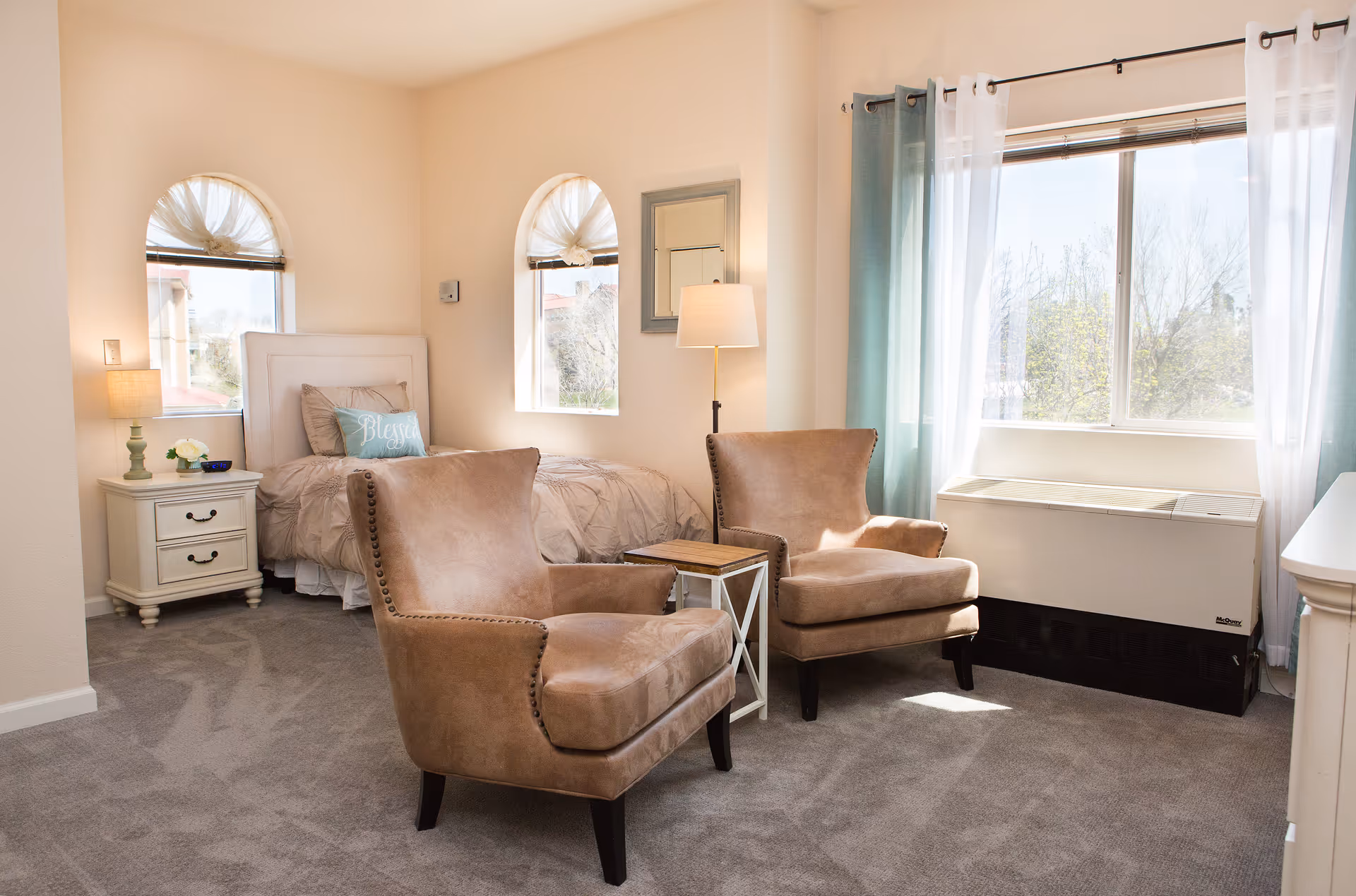A bright and cozy assisted living bedroom featuring a single bed with beige bedding and a decorative pillow that says 'Blessed'. There are two arched windows with sheer curtains behind the bed, a nightstand with a lamp and flowers, two tan upholstered armchairs with a small wooden side table between them, a floor lamp, and a large window with sheer white and teal curtains letting in natural light.