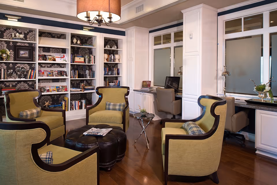 Cozy lounge with green upholstered armchairs arranged around a leather ottoman in front of built-in bookshelves and computer workstations.