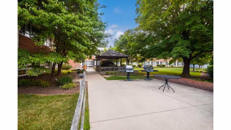 Outdoor courtyard area with a concrete walkway, two barbecue grills, a round table, green grass, trees, and a gazebo in the background surrounded by a multi-story building.