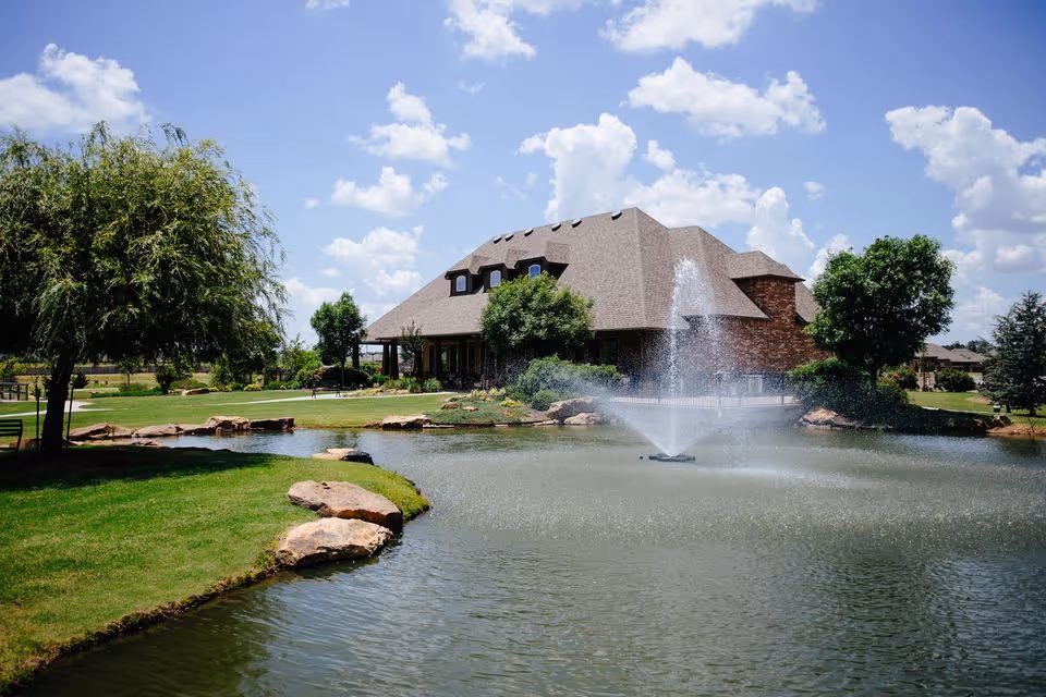 Clubhouse-style building beside a pond with a central fountain, grassy lawns, and trees under a blue sky.
