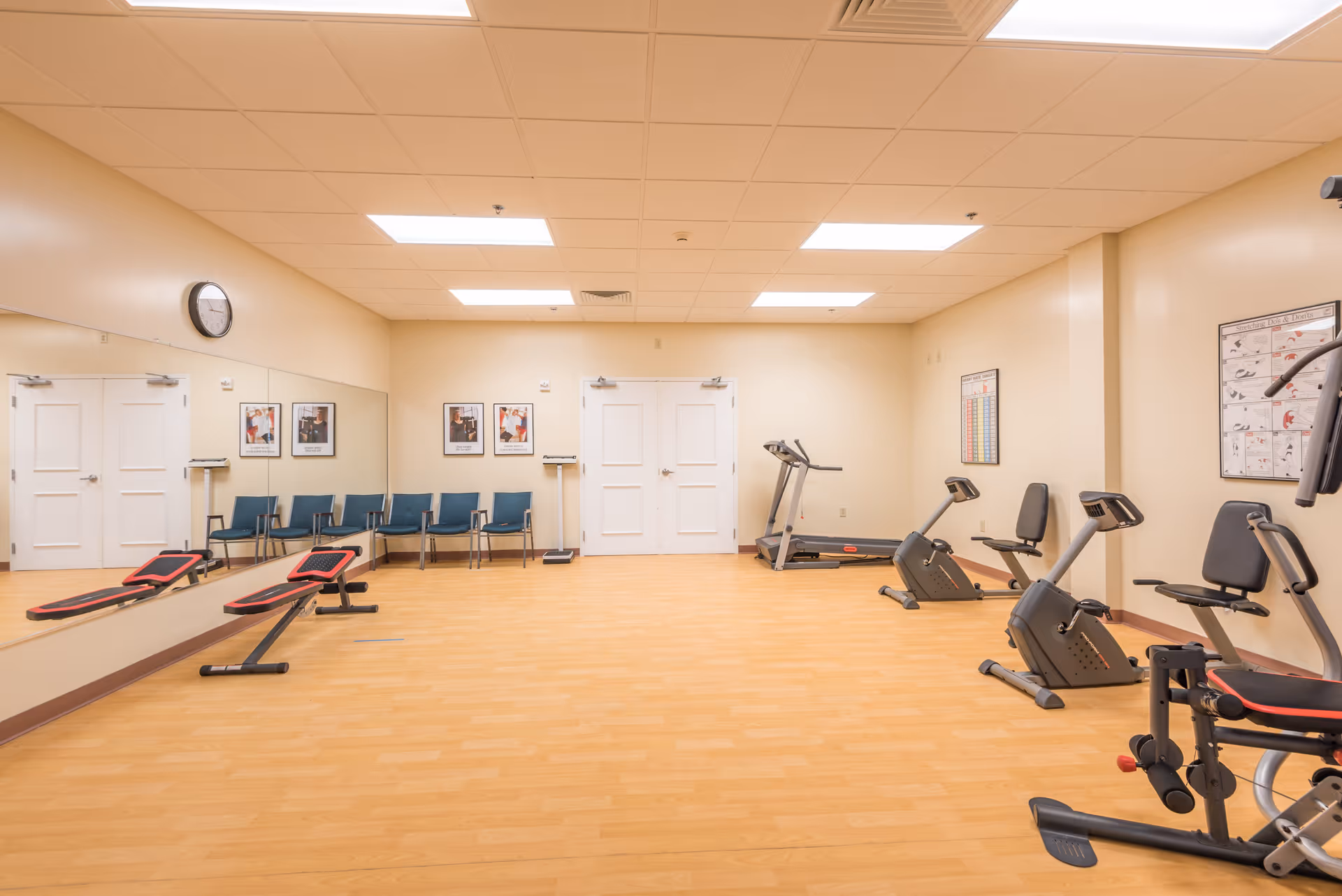 A bright fitness room with treadmills, stationary bikes, benches, a row of chairs along a mirrored wall, and exercise charts on the walls.
