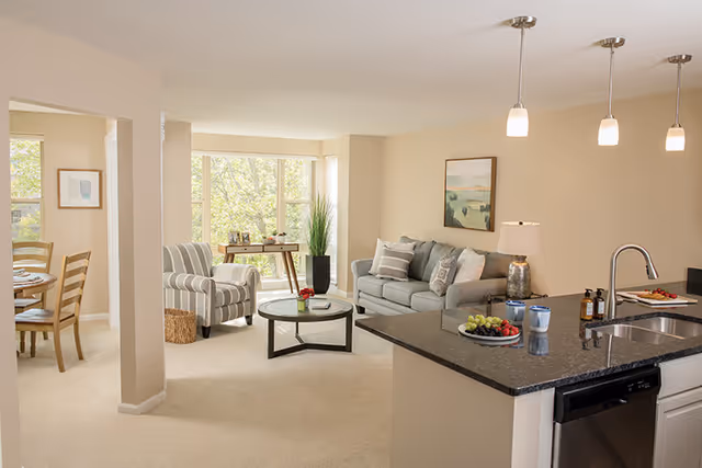 Bright and spacious senior living community interior featuring a living room with a gray sofa, striped armchair, round glass coffee table, and a side table with a lamp. Adjacent to the living room is a kitchen area with a black granite countertop island, sink, and dishwasher. In the background, a dining area with a wooden table and chairs is visible near large windows letting in natural light.