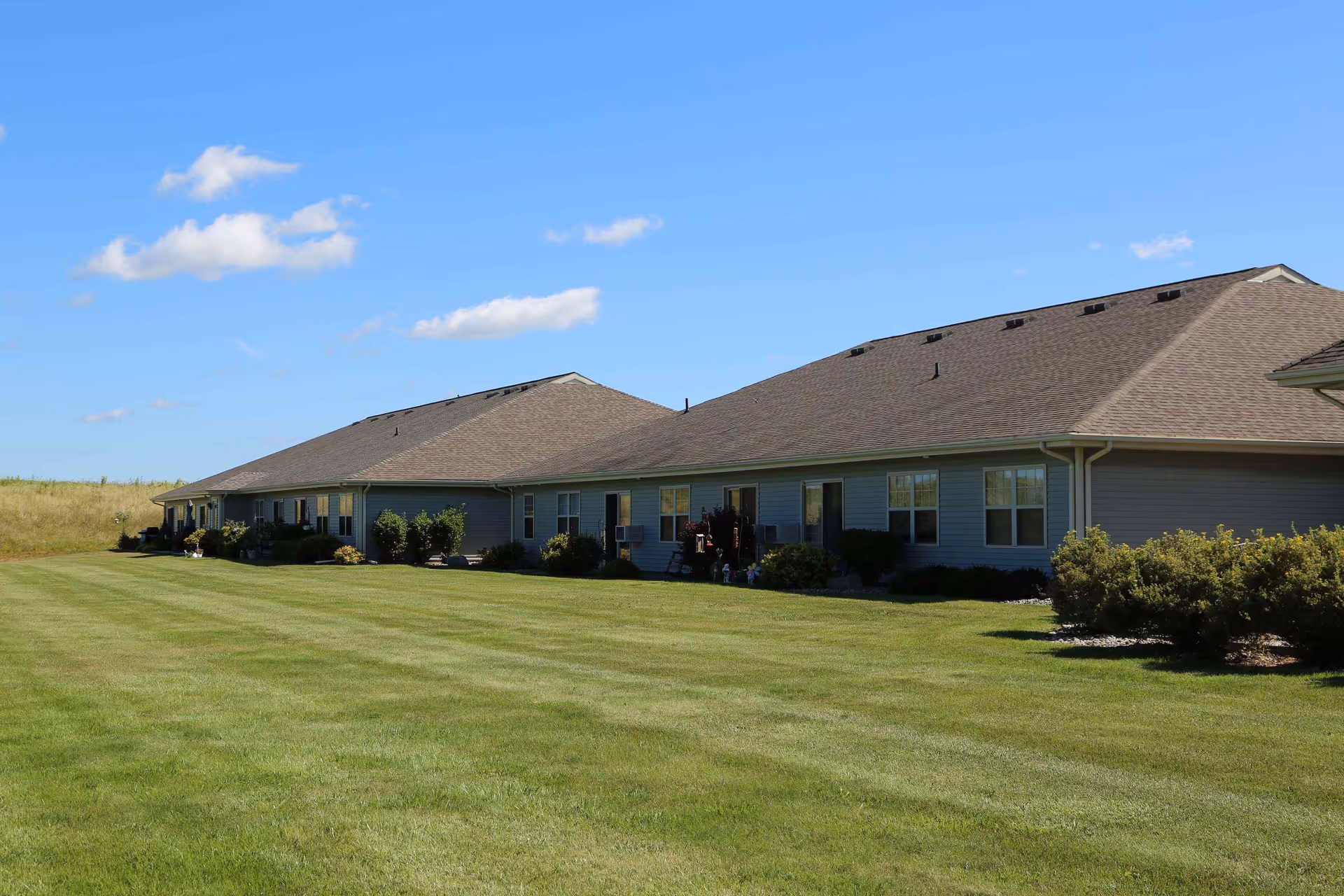 Exterior view of a single-story senior living facility building with gray siding and brown shingled roofs, surrounded by a well-maintained green lawn and some bushes under a blue sky with a few clouds.