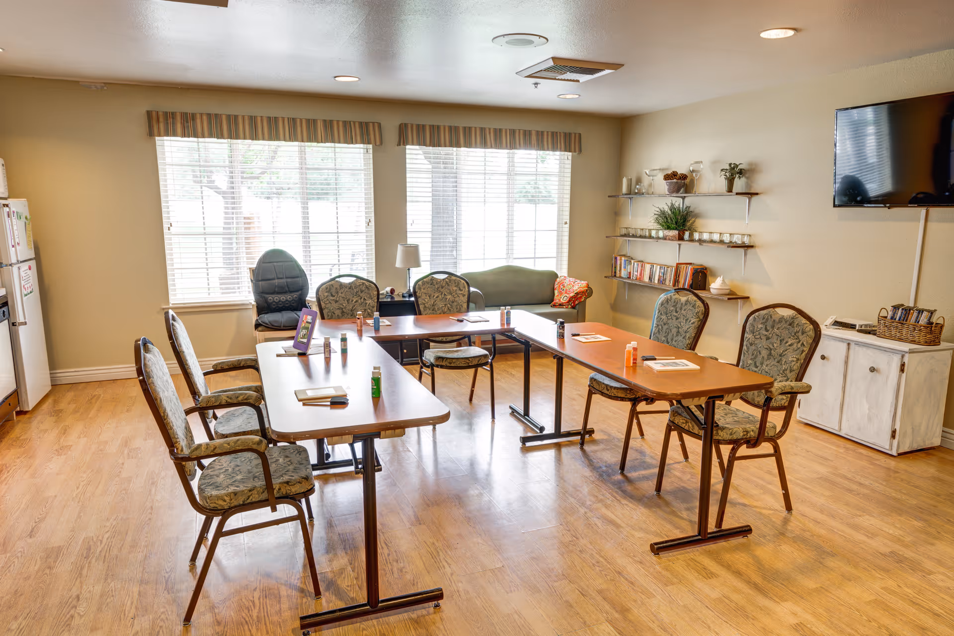 A bright room with wooden floors and large windows with blinds. There are two rectangular tables arranged in a U-shape with six cushioned chairs around them. On the tables are small bottles and papers. Against the wall is a green cushioned bench, a lamp, and a TV mounted on the wall. Shelves with books, plants, and decorative items are also visible.