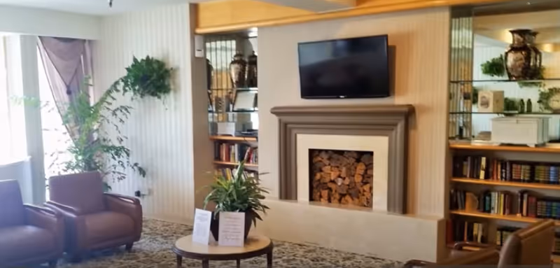 A cozy living room area with two brown leather armchairs, a round wooden coffee table with a plant and a small sign on it, a fireplace filled with stacked firewood, a flat-screen TV mounted above the fireplace, built-in wooden bookshelves with books and decorative items, and a large window with sheer curtains letting in natural light.
