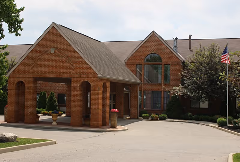 Front exterior view of a senior living facility with a brick entrance canopy, large windows, landscaping with bushes and trees, and an American flag on a flagpole.