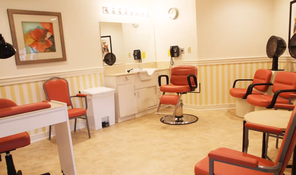 Interior of a senior living facility salon with red cushioned chairs, a hair washing station, a manicure table, a wall-mounted phone, a large mirror, and framed artwork on the wall. The room has yellow and white striped wallpaper and beige flooring.