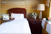A neatly made single bed with white bedding and a wooden headboard next to a wooden nightstand. The nightstand holds a white lamp with a cylindrical shade and a white flower arrangement. A small framed picture and a reed diffuser are placed on the bed's headboard. The room has warm yellow walls and a window with curtains partially visible.