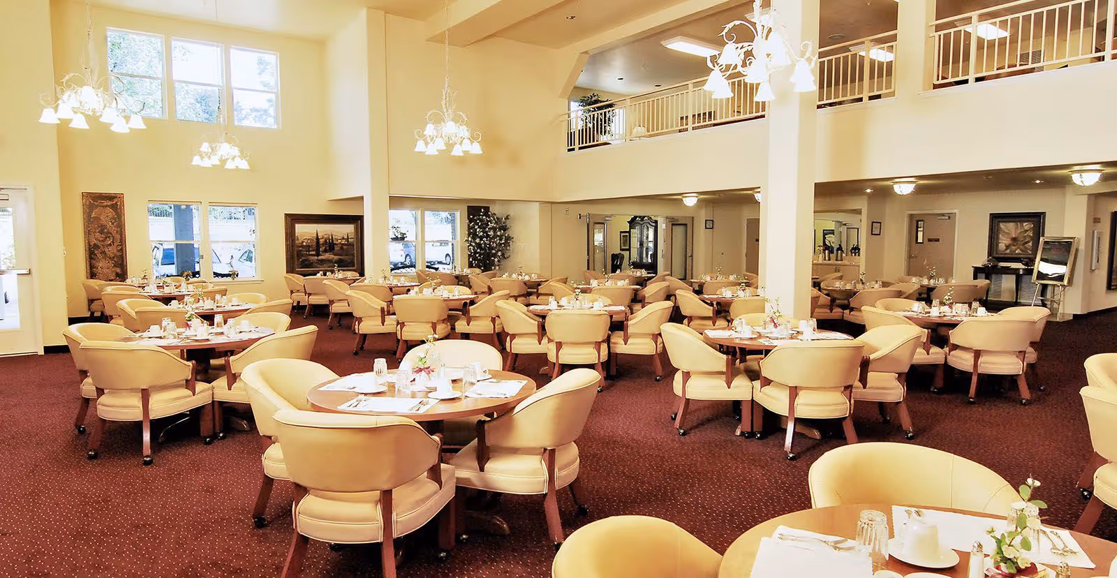 Spacious dining room with multiple round tables set with white tableware and beige cushioned chairs. The room has high ceilings with hanging chandeliers, large windows letting in natural light, and decorative plants and paintings on the walls.