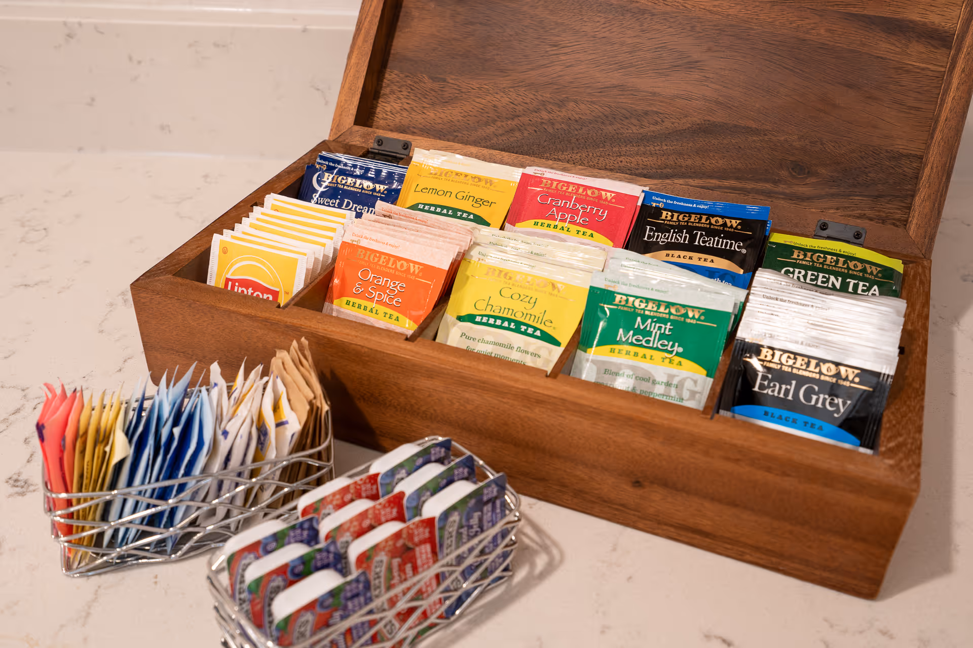 Wooden tea box on a countertop displaying assorted individually wrapped tea bags with nearby wire baskets holding sweetener packets and mints.