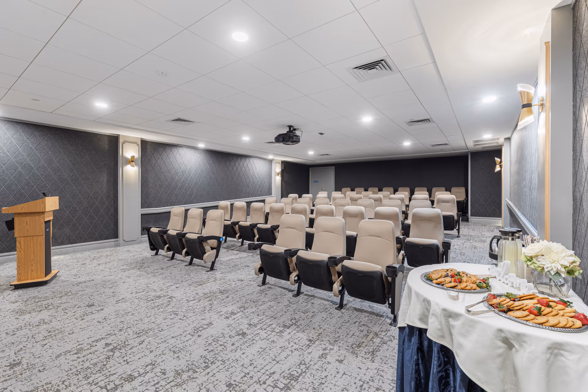 Small presentation/theater room with rows of beige auditorium seats, a podium, ceiling projector, and a refreshment table with trays of snacks.
