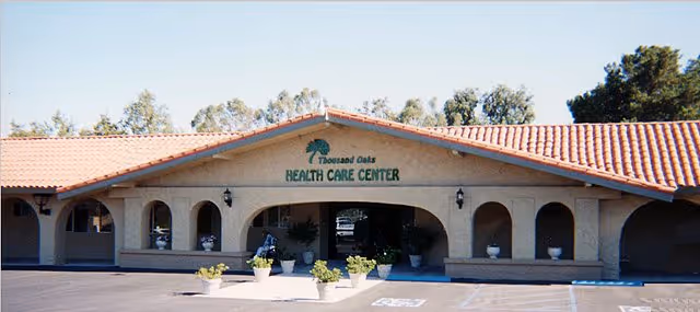 Front exterior view of Thousand Oaks Health Care Center building with a tiled roof, arched windows and doorways, potted plants near the entrance, and a parking area in front.