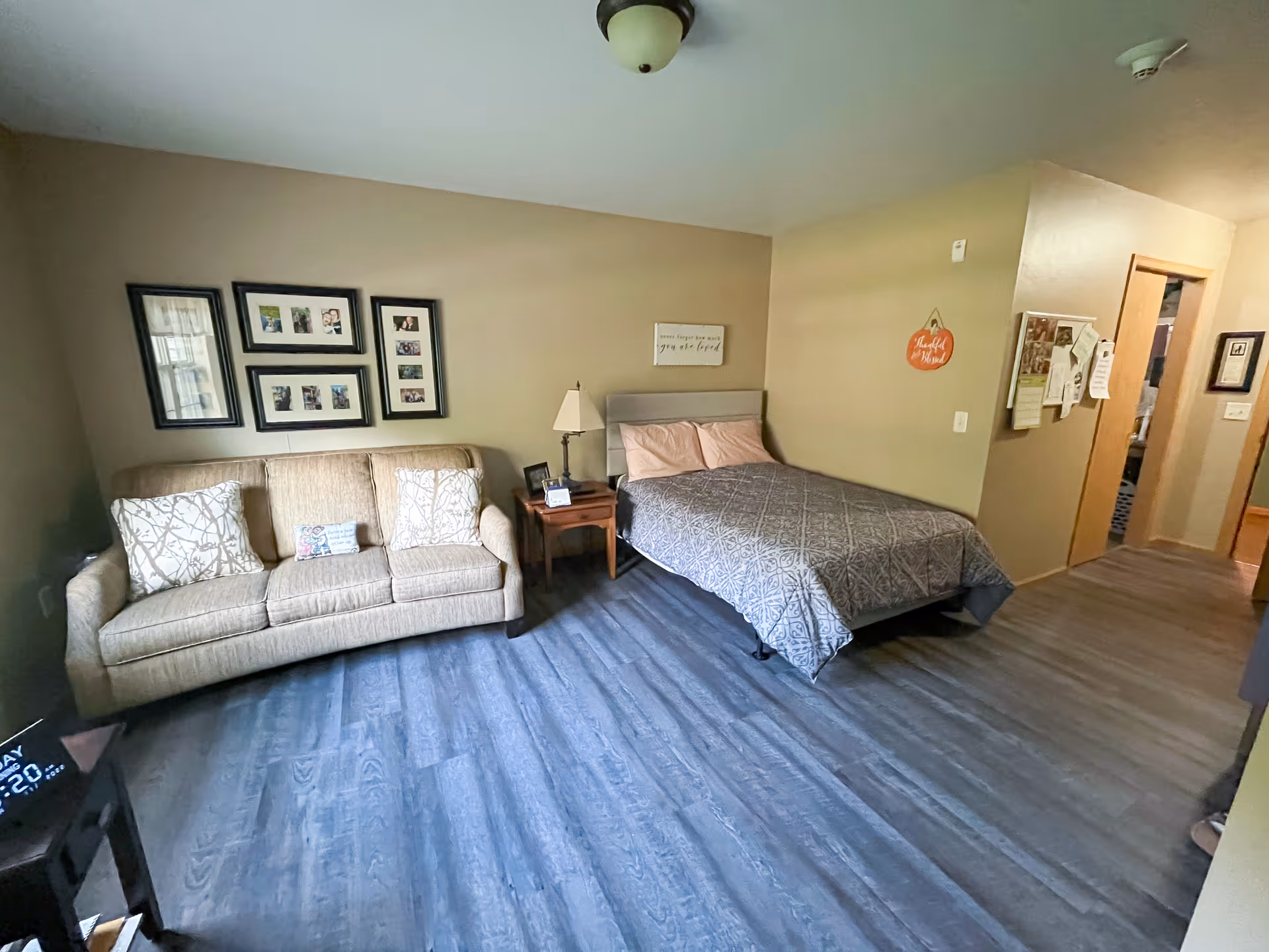 A cozy bedroom in a senior living facility featuring a bed with a gray patterned comforter and two pillows, a beige sofa with decorative pillows, a wooden nightstand with a lamp and picture frame, and framed photos on the wall. The room has wood-look flooring and beige walls with a small decorative sign hanging near the doorway.