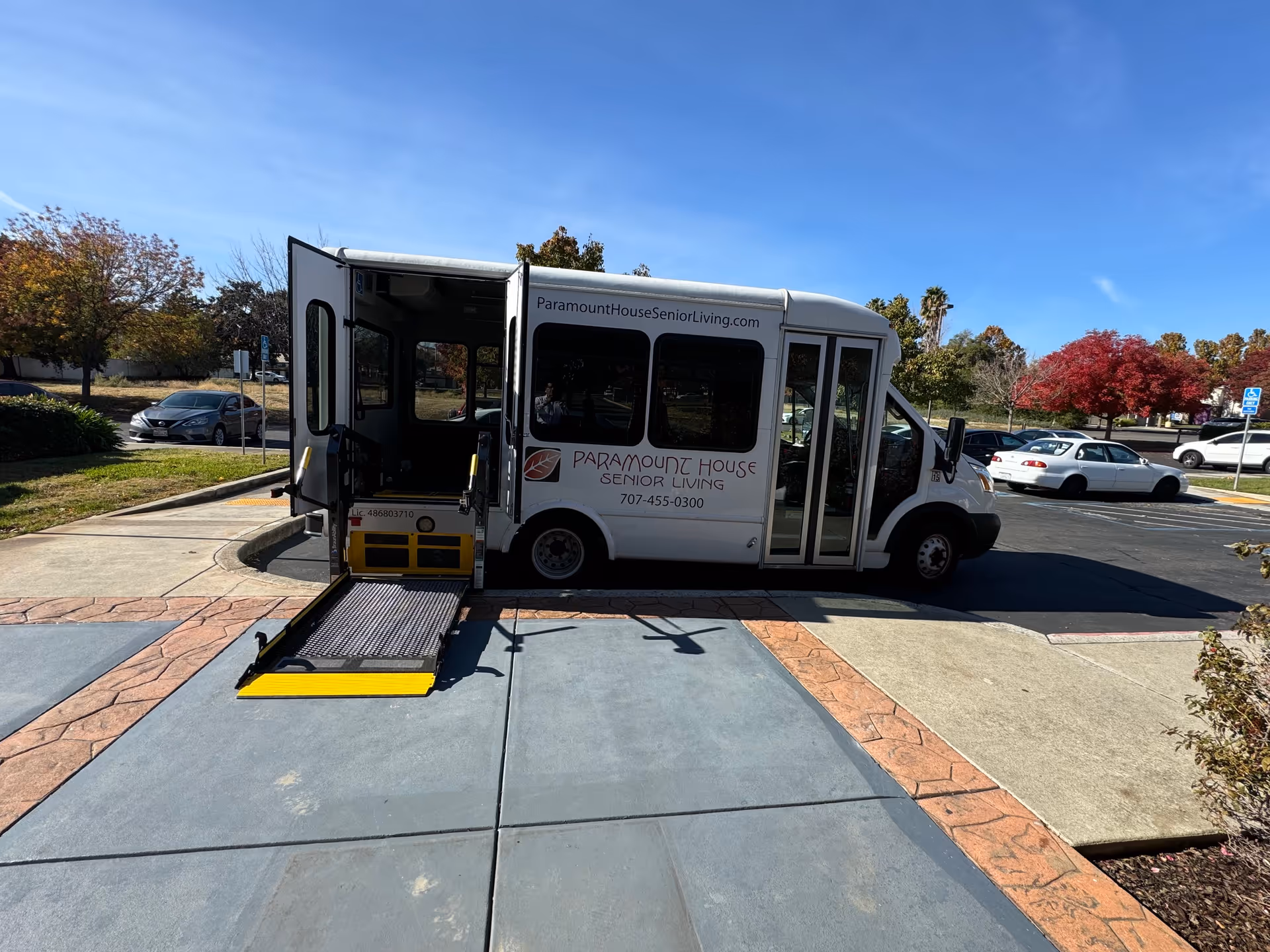 A white shuttle bus with an extended wheelchair ramp parked on a driveway near a sidewalk. The bus has the text 'Paramount House Senior Living' along with a phone number and website on its side. The background shows a parking lot with several cars and trees with autumn foliage under a clear blue sky.
