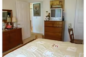 A bedroom with a bed covered in a light-colored quilt with patterns, a wooden dresser with a TV and decorative items on top, a wooden chair, and an open door leading to another room with a framed picture on the wall.