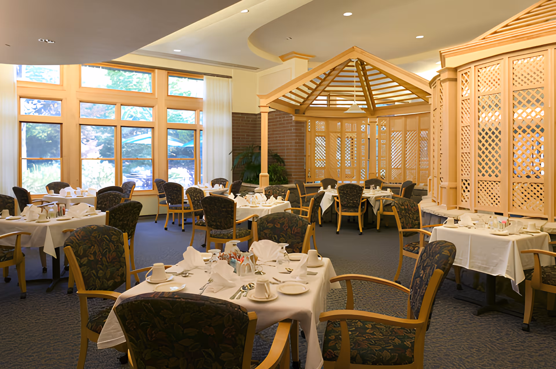 A bright dining room in a senior living community with multiple tables covered in white tablecloths, set with cups, plates, napkins, and silverware. The room features large windows letting in natural light, patterned upholstered chairs, and a wooden lattice structure in the background.