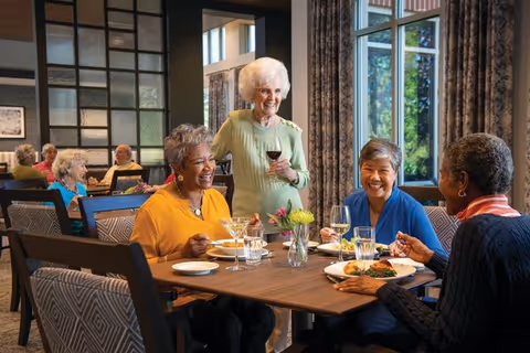 Four elderly women enjoying a meal together at a wooden dining table in a well-lit dining room with large windows and patterned chairs. One woman is standing and holding a glass of red wine, while the other three are seated and smiling, with plates of food and drinks in front of them.