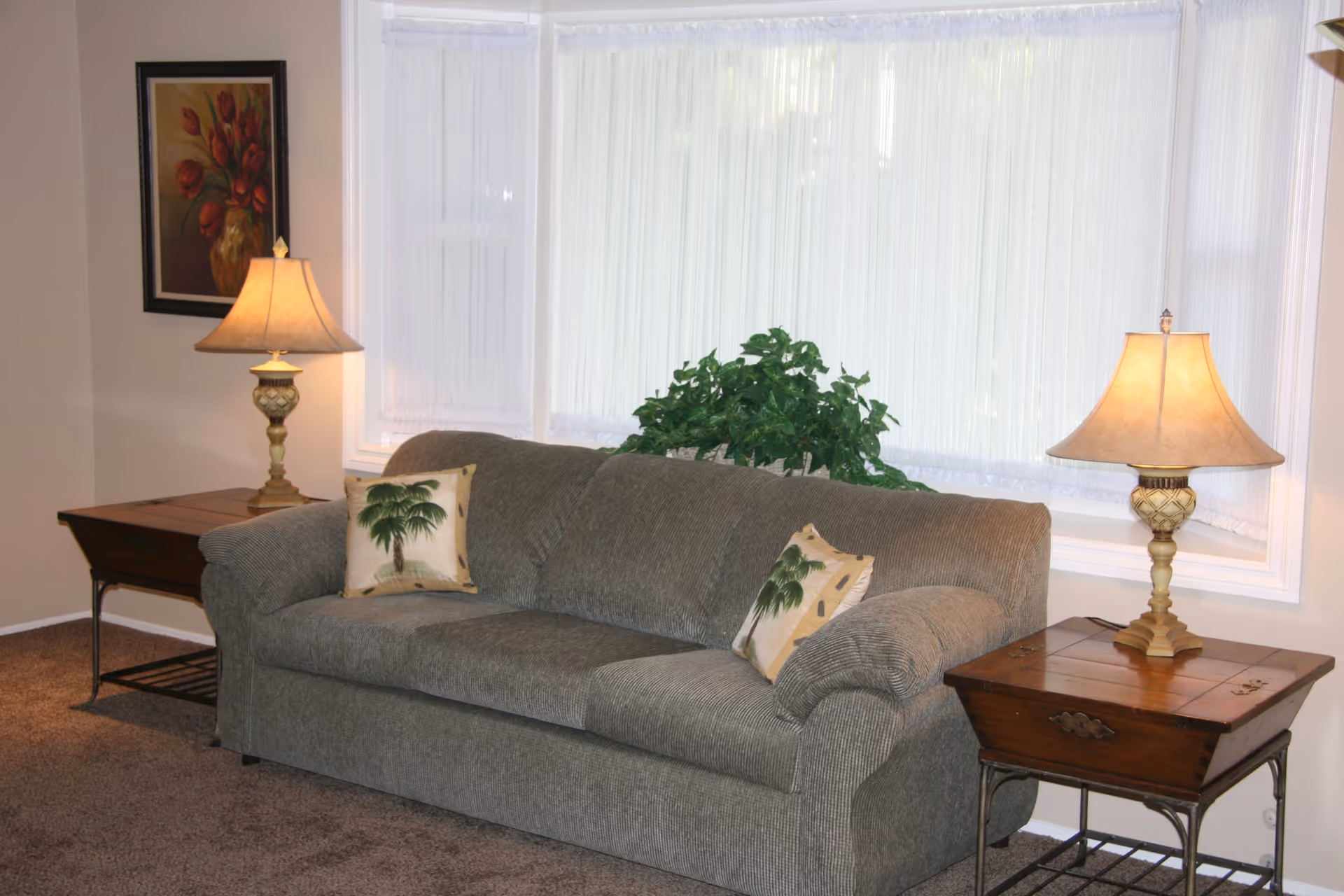 A cozy living room area with a gray upholstered sofa adorned with two decorative pillows featuring palm tree designs. On either side of the sofa are wooden side tables, each with a classic table lamp emitting warm light. Behind the sofa is a large window with sheer white curtains, and a green leafy plant is placed in front of the window. A framed painting of red flowers in a vase hangs on the wall to the left.