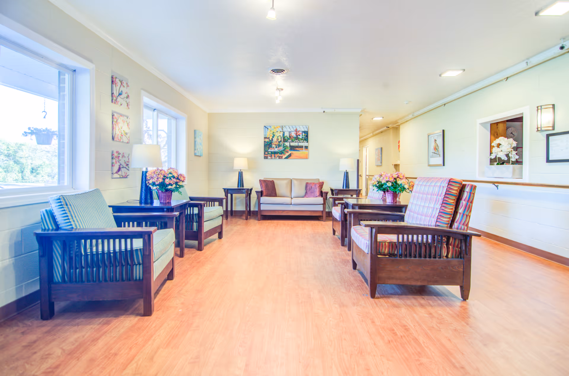 A bright and spacious senior living facility common area with wooden flooring, multiple armchairs with patterned cushions, a beige sofa with pillows, side tables with lamps and flower arrangements, wall art, and large windows letting in natural light.