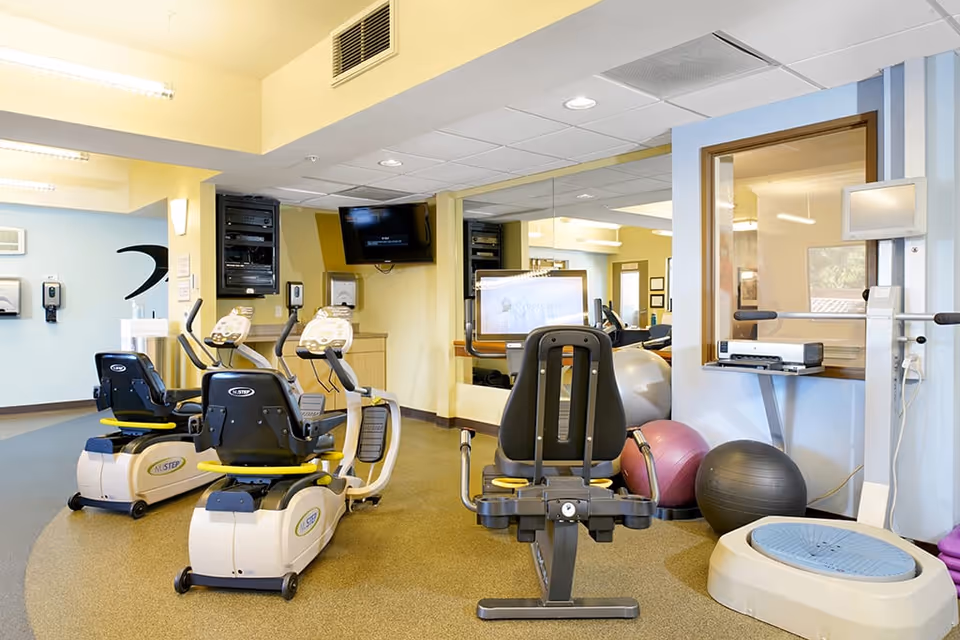 A well-lit exercise room with stationary recumbent bikes, exercise balls, a balance platform, and a wall-mounted television. The room has light-colored walls, a large mirror, and fitness equipment arranged neatly on a carpeted floor.
