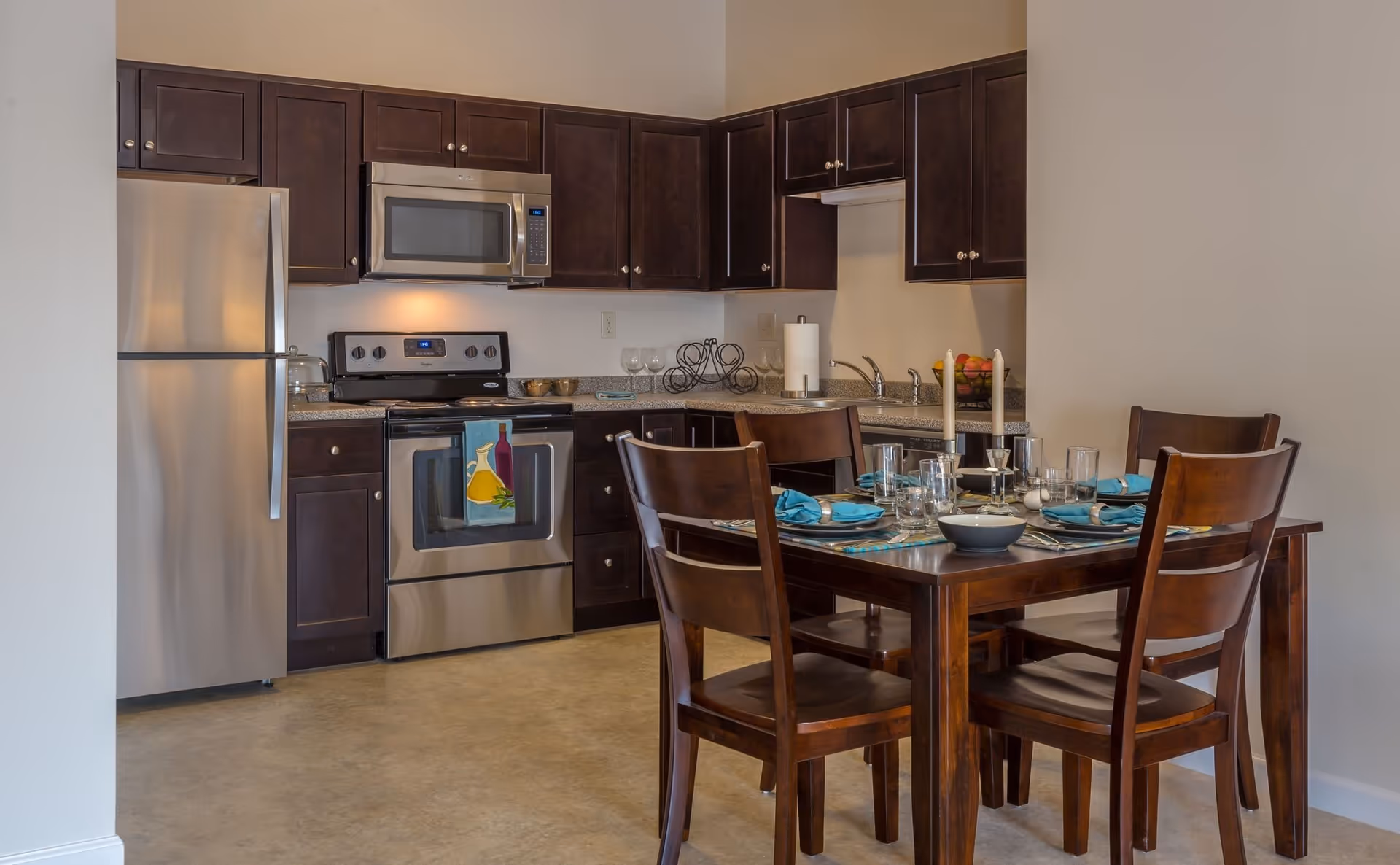 A modern kitchen and dining area featuring dark wood cabinets, stainless steel refrigerator, stove, and microwave. The dining table is set with blue napkins, glassware, and candles, with four wooden chairs surrounding it.